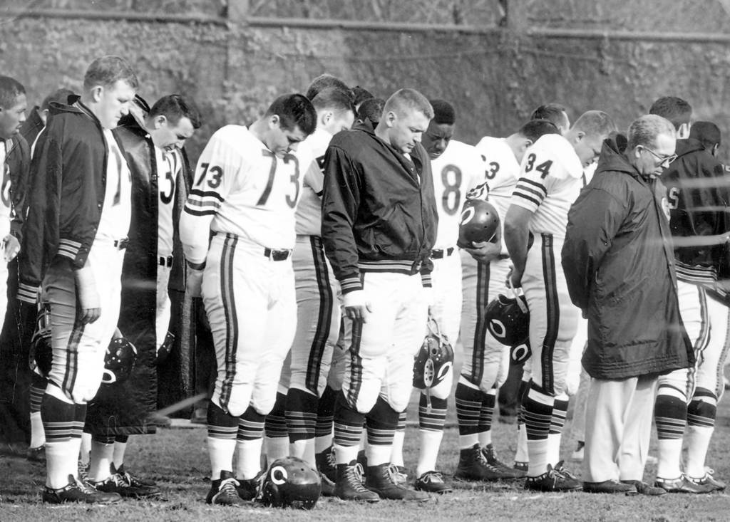 Chicago Bears bench before the beginning of a game against the Pittsburgh Steelers on Nov, 25, 1963. The Bears bow their heads in memory of President John F. Kennedy. From left are: Bennie McRae (26), Bob Kilcullen, Joe Fortunato, Steve Barnett (73), Wetoska, Willie Galimore (28), Joe Marconi (34), and coach Phil Handler.
