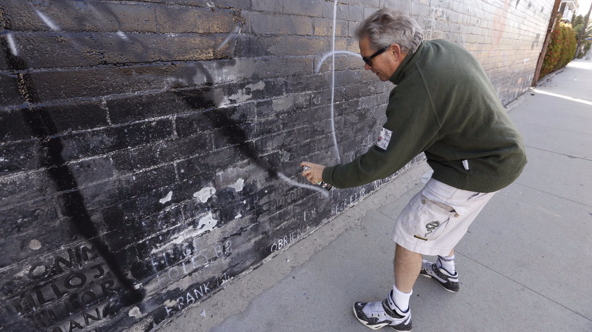 Steven Barber uses black spray paint to cover the white letters of graffiti on the wall.